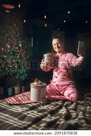 Cheerful cute funny man in pink sleepwear with Christmas present making selfie photo and sit on the bed near decorated fir tree