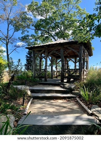 A gazebo made out of wood and natural logs in the middle of the wilderness.  There are benches, wide steps, and lots of shade in this relaxing area.