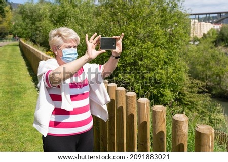 Senior woman with face mask doing panoramic video with cellphone at park on sunny day. Elder lady standing by fence taking photo with smart phone on natural landscape