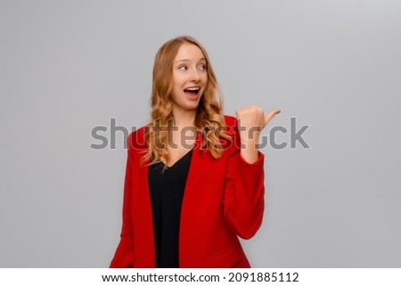Hey look there. Smiling excited blonde young woman pointing finger to the right, showing advertisement, demonstrating promo offer, standing in red blazer against gray background