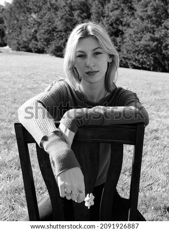 black and white image of a young woman facing the camera sitting relaxed in a chair outdoors with a small flower in her hand