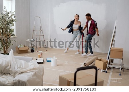 Entertaining cheerful friends spend time together, have fun while renovating the room, apartment, hold paint rollers in their hands, paint walls with white paint dancing singing joy of buying flat Royalty-Free Stock Photo #2091958183