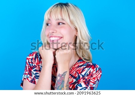 Caucasian girl wearing floral dress isolated over blue background holds hands under chin, glad to hear heartwarming words from stranger