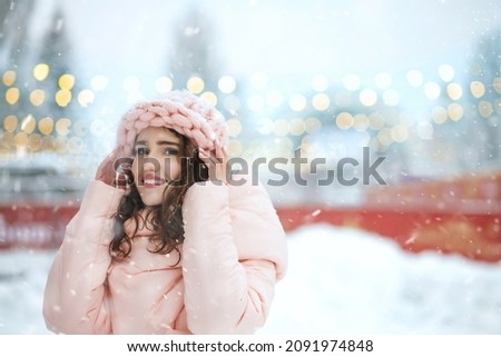 Charming young woman wears knitted cap walking at the holiday fair against the background of garlands. Empty space