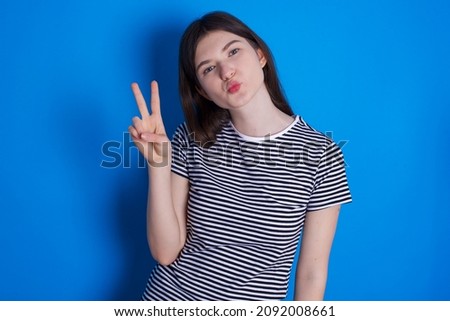 Caucasian woman wearing striped T-shirt isolated over blue background makes peace gesture keeps lips folded shows v sign. Body language concept