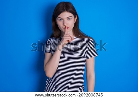 Surprised Caucasian woman wearing striped T-shirt isolated over blue background makes silence gesture, keeps finger over lips and looks mysterious at camera