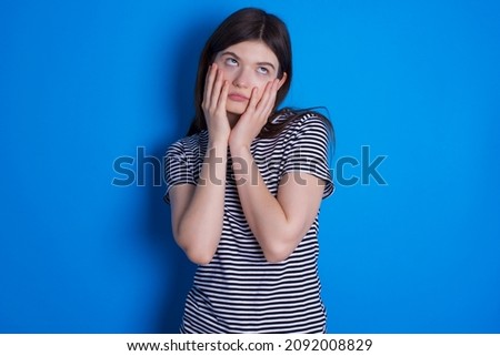 Caucasian woman wearing striped T-shirt isolated over blue background keeps hands on cheeks has bored displeased expression. Stressed hopeless model