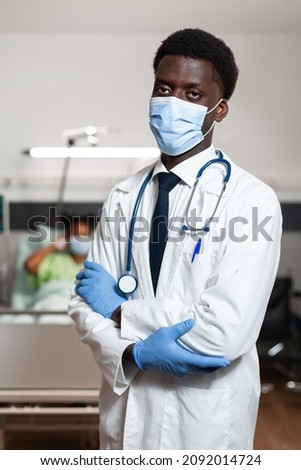 Portrait of african american practitioner doctor with protective face mask to prevent infection with covid19 looking into camera while standing in hospital ward. In background patient recovering