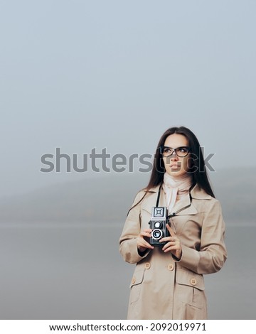 Retro Woman Holding a Vintage Camera Wearing Trench Coat. Female detective wearing cat eye eyeglasses and 70s inspired fashion items
