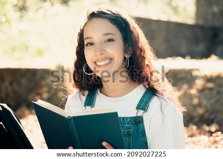 Young beautiful arab curly student girl sitting outdoors reading a book and taking notes in notebook. Online and remote education concept. In a campus at sunset with a warm light. Copy space for text