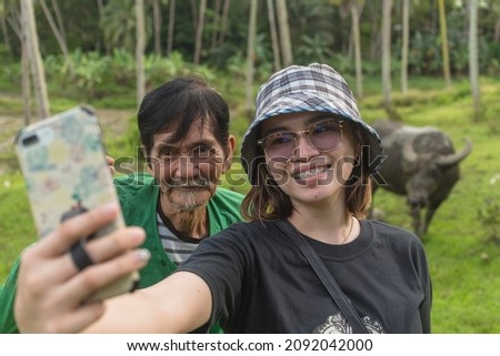 A young modern asian woman takes a selfie with her farmer dad while at a rice paddy. Bonding during a vacation at the countryside.