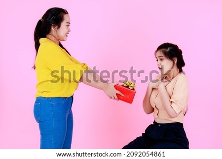 Studio shot of Asian chubby female model in casual wear giving red Birthday Christmas New Year present gift box with gold ribbon to friend or sister who sitting surprising exciting on pink background.