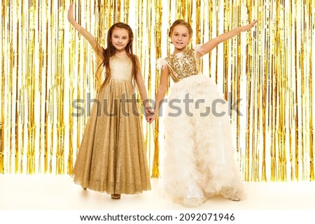 Kid's fashion and holidays. Beautiful little girls in gorgeous evening dresses pose on the background with shiny gold foil curtains. Children's party. Full length shot.