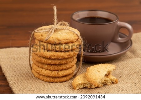 Stack of oatmeal cookies with sesame seeds tied with string on burlap cloth on wooden table. Cup of coffee. Front horizontal view.