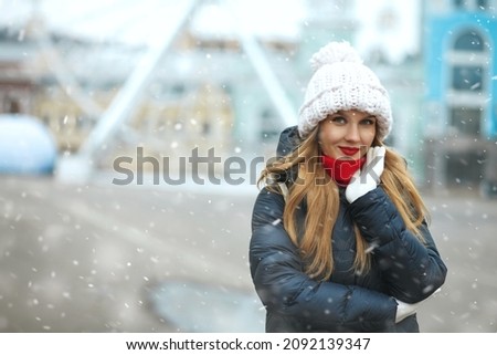 Elegant blond woman wearing knitted cap walking at the city during snowfall. Space for text