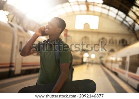 Young man waiting for the train standing at the railway station. Happy guy talking to the phone while waiting for the train