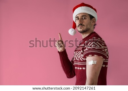 Christmas vaccination campaing. Smiling young man with santa hat and red sweater with bandage in arm points away over pink background