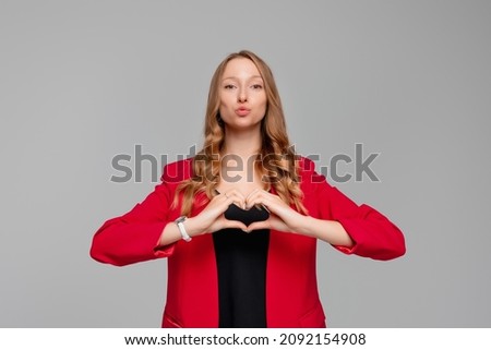 My heart belongs to you. Beautiful woman shows heart symbol, shapes love sign with hands, wears red blazer, gray background. Be my Valentine