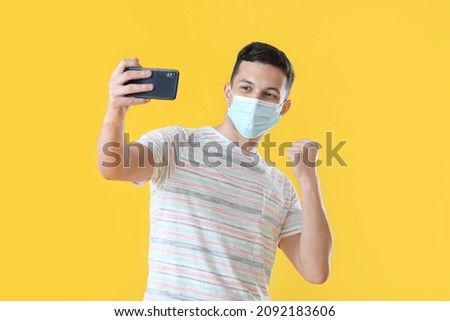 Young man in medical mask taking selfie on color background