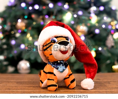 a toy tiger in a santa claus hat on the background of a christmas tree