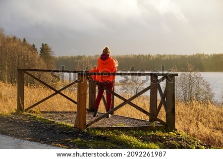 Woman in nature takes photo on sunset. standing with his back in a red jacket. Autumn lake and wooden viewpoint. Rainy. High quality photo