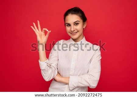 Photo of young positive happy beautiful brunette woman with sincere emotions wearing white shirt isolated over red background with copy space and showing ok gesture. It's fine