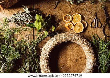 DIY Christmas wreath making on wooden tabletop with evergreen tree branches dry fruits oranges pine cones and scissors, top view