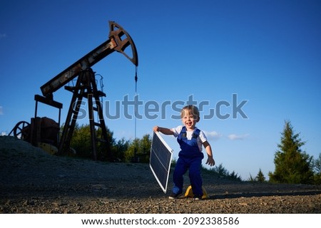 Young kid engineer having fun outdoors in field with oil pump jack at sunny summer day. Smiling child holding small solar panel. Alternative fotovoltaic ecoenergy conception.