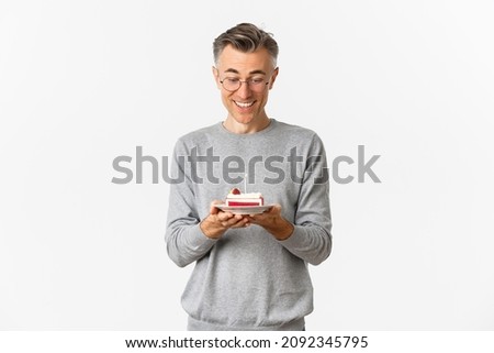 Image of happy, attractive middle-aged man in glasses and grey sweater, celebrating his birthday, smiling and looking at b-day cake, making a wish, standing over white background