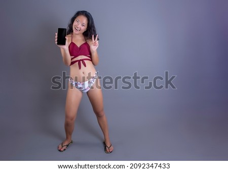 beautiful young asian girl in bikini showing smart phone screen and making victory sign with her fingers