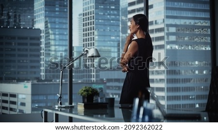 Successful Businesswoman in Stylish Dress Looking out of the Window at Big City in Downtown Area. Confident Female CEO Working on Financial Projects. Manager at Work Planning Marketing Campaign.