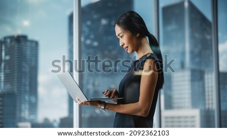 Beautiful Portrait of an Asian Businesswoman in Stylish Black Dress Using Laptop Computer, Posing Next to Window in City Office. Confident Female CEO Smiling. Successful Diverse Business Manager.