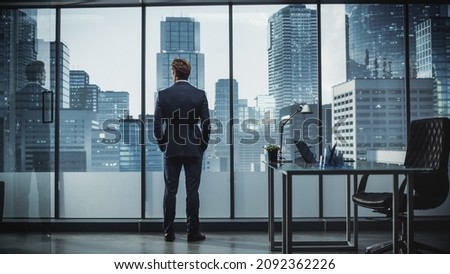 Successful Young Businessman in a Perfect Tailored Suit Standing in His Modern Office Looking out of the Window on Big City with Skyscrapers. Successful Finance Manager Planning Project Strategy. Royalty-Free Stock Photo #2092362226