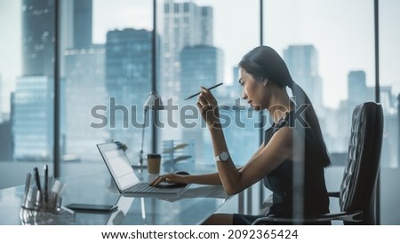 Successful Businesswoman in Stylish Dress Sitting at a Desk in Modern Office, Using Laptop Computer, Next to Window with Big City View. Successful Finance Manager Planning Work Projects. Royalty-Free Stock Photo #2092365424