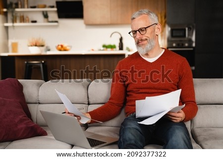 Troubled caucasian serious mature businessman freelancer working remotely on laptop, doing difficult paperwork, multitasking, solving problems remotely at home Royalty-Free Stock Photo #2092377322