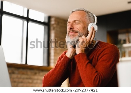 Relaxed mature middle-aged man businessman freelancer listening to the music, podcast, e-book, radio programme, playlist, musical track in headphones while working remotely on laptop at home