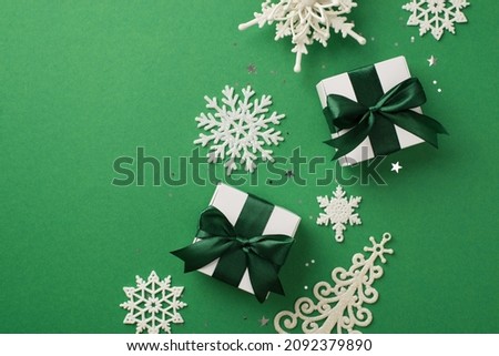Top view photo of christmas decorations white snowflake pine ornaments star shaped confetti sequins and white gift boxes with green ribbon bows on isolated green background with copyspace