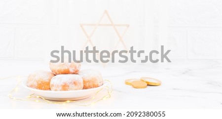 Donut with powdered sugar and cream in a saucer with a garland, coins and wooden Star of David lie on a white wooden table, close-up side view. Hanukkah celebration concept.