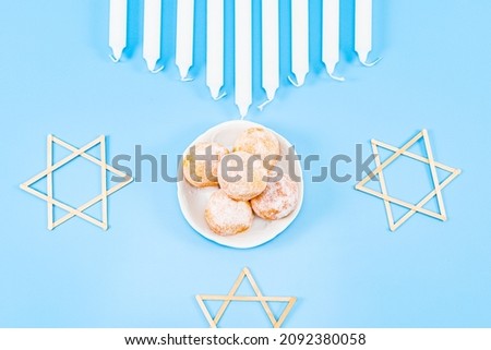 Small donuts with powder in a saucer, nine white candles and three wooden stars of David lie on a blue background, flat lay close-up. Hanukkah Celebration Concept.