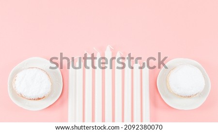 Nine white candles and two donuts with powdered sugar in saucers lie on a pink background with copy space, close-up. Hanukkah celebration concept.
