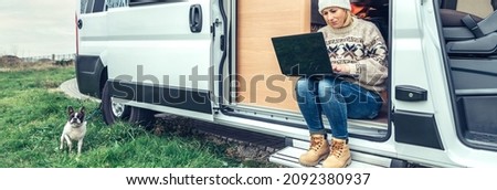 Woman teleworking sitting in the door of a camper van while her partner works inside