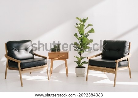 Home decor on side table with drawer standing near two leather armchairs and potted house plants. Recreation, relaxation concept. Wooden table and office chairs in bright copy space living room Royalty-Free Stock Photo #2092384363