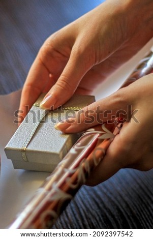 Close-up of female hands holding a small silver gift box and wrapping this box with ribbon in golden wrapping paper. Moment of preparing a present handmade. Vertical photo. Image with selective focus