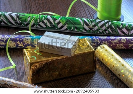 Two gift boxes in gold and silver wrapping stand on top of each othe. In the background rolls of colored wrapping paper with curled green decorative wrapping tape on them. Image with selective focus