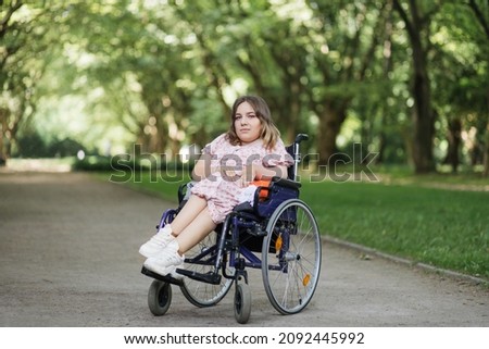 Portrait of pleasant young woman with spinal muscular atrophy smiling on camera among green summer park. Female person who using wheelchair. Concept of people with disability. Royalty-Free Stock Photo #2092445992