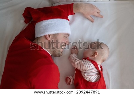Father with his baby girl wearing Santa hats resting on bed.