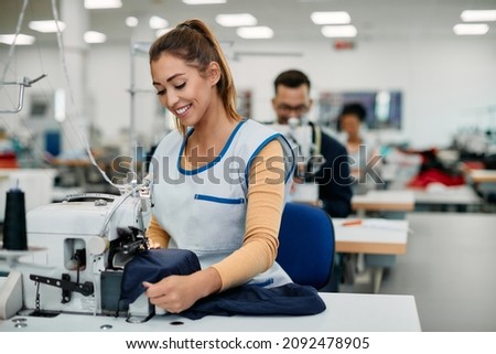 Young happy seamstress working in textile factory and sewing fabric. Royalty-Free Stock Photo #2092478905