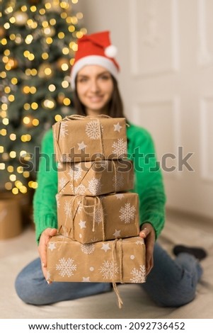 The young smiling girl holds brown gifts near Christmas festive tree. Happy woman with a red hat on head and green sweater see in a camera with boxes in focus