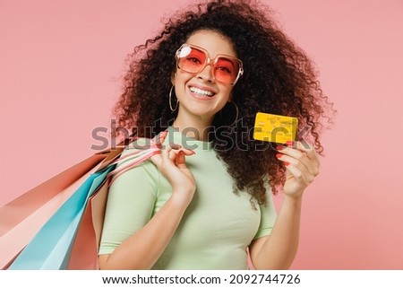 Side view profile young curly latin woman 20s wears mint t-shirt sunglasses hold package bags with purchases after shopping credit bank card isolated plain pastel light pink background studio portrait