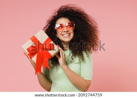 Happy young curly latin woman 20s wears mint t-shirt sunglasses hold shake red present box with gift ribbon bow try to guess what inside isolated on plain pastel light pink background studio portrait